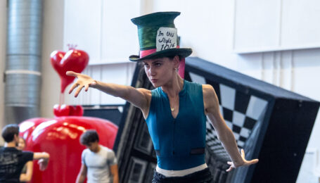 In a blue leotard, black pants, and the Mad Hatter's oversized top hat, Amelia Townsend points dramatically at something off camera.
