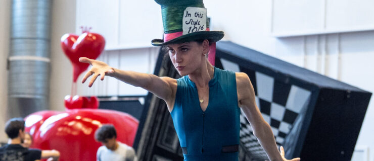 In a blue leotard, black pants, and the Mad Hatter's oversized top hat, Amelia Townsend points dramatically at something off camera.