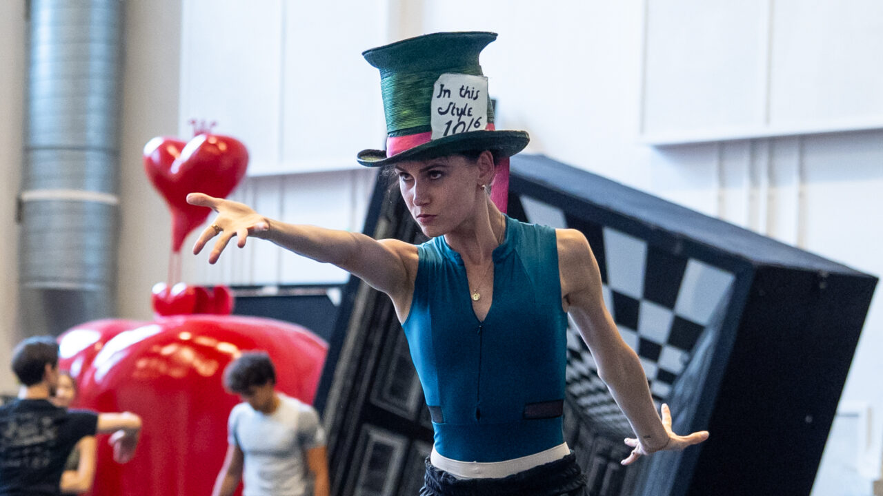 In a blue leotard, black pants, and the Mad Hatter's oversized top hat, Amelia Townsend points dramatically at something off camera.