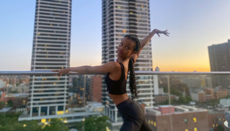 Murray—a Black woman wearing a black crop top and black pants, her long braids pulled into a low ponytail—poses on a city rooftop. Her left arm drapes elegantly over the railing as she extends her right arm up and out behind her.