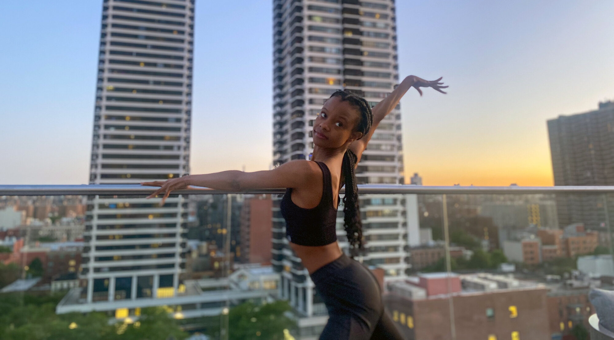 Murray—a Black woman wearing a black crop top and black pants, her long braids pulled into a low ponytail—poses on a city rooftop. Her left arm drapes elegantly over the railing as she extends her right arm up and out behind her.