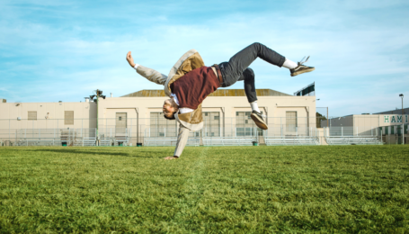 A male dancer outside on the grass in front of a school building. He flips on one hand with the rest of his body levitating in the air.