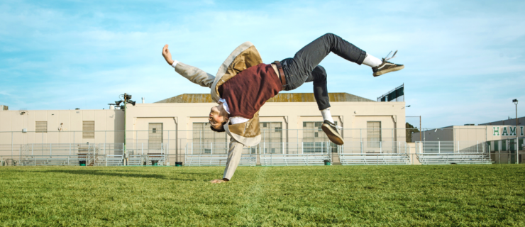 A male dancer outside on the grass in front of a school building. He flips on one hand with the rest of his body levitating in the air.