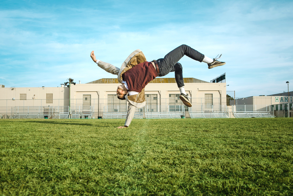 A male dancer outside on the grass in front of a school building. He flips on one hand with the rest of his body levitating in the air.