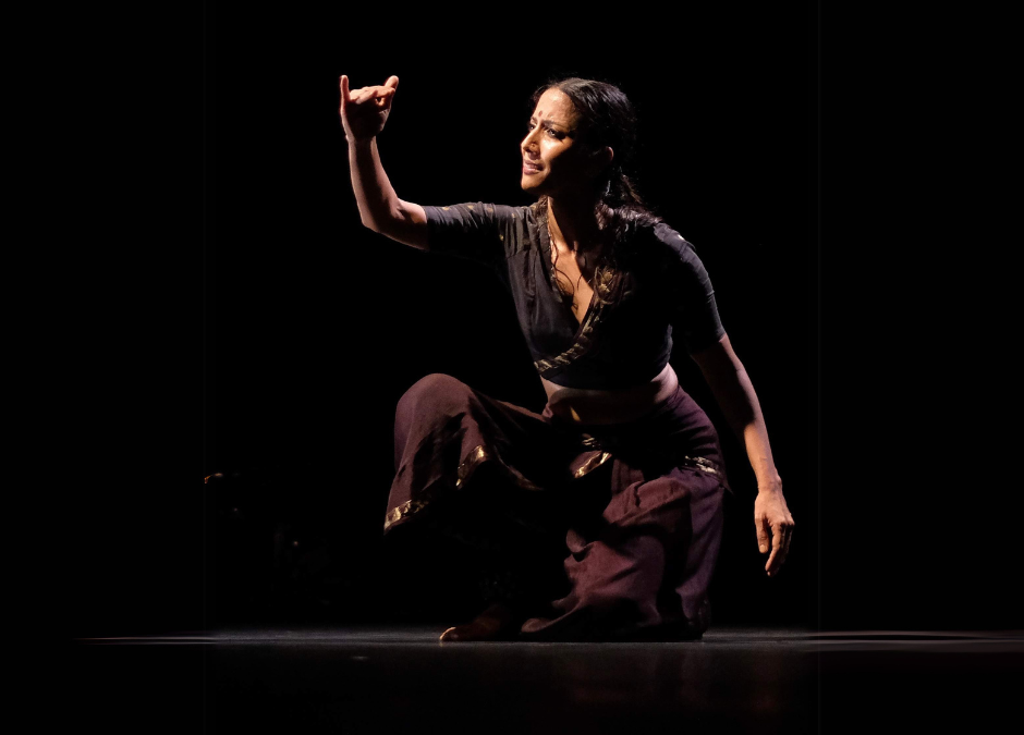 A Bharatanatyam dancer kneeling on the floor looking at her hand lifted in front of her face