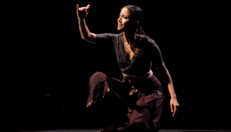 A Bharatanatyam dancer kneeling on the floor looking at her hand lifted in front of her face