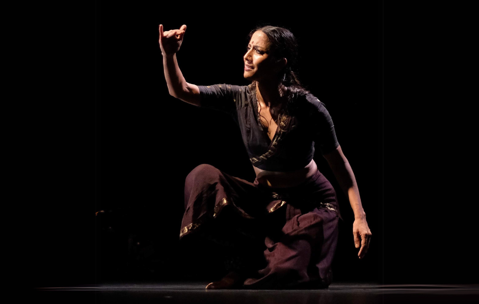 A Bharatanatyam dancer kneeling on the floor looking at her hand lifted in front of her face