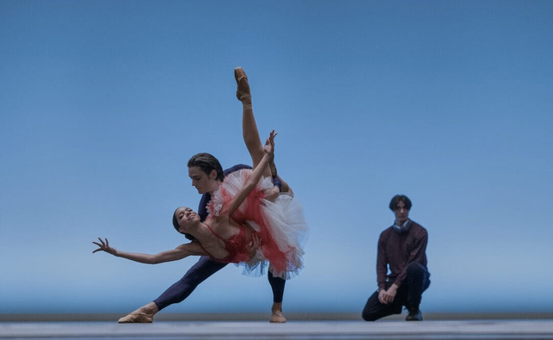 Onstage in front of a light blue backdrop, two dancers do a triumphant "fish" pose, the supporting partner lunging to the side and holding the other dancer in a dipped position with one leg tucked and the other extended long. Behind them, a choreographer crouches and watches with hands on his lap.