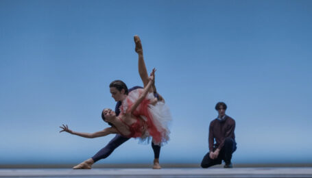 Onstage in front of a light blue backdrop, two dancers do a triumphant "fish" pose, the supporting partner lunging to the side and holding the other dancer in a dipped position with one leg tucked and the other extended long. Behind them, a choreographer crouches and watches with hands on his lap.