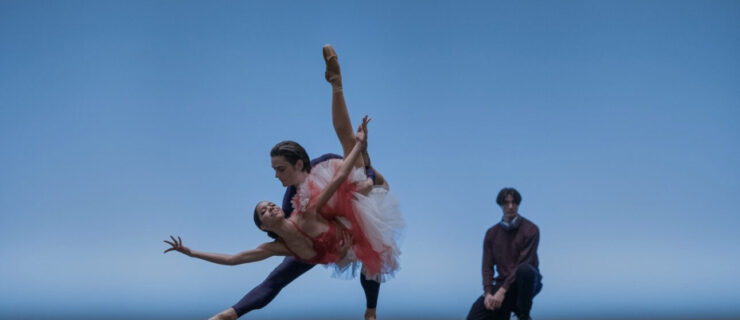 Onstage in front of a light blue backdrop, two dancers do a triumphant "fish" pose, the supporting partner lunging to the side and holding the other dancer in a dipped position with one leg tucked and the other extended long. Behind them, a choreographer crouches and watches with hands on his lap.