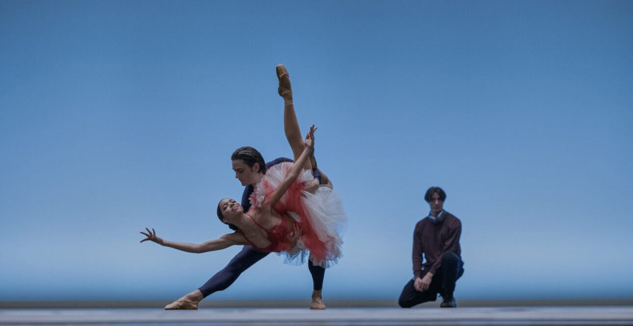 Onstage in front of a light blue backdrop, two dancers do a triumphant "fish" pose, the supporting partner lunging to the side and holding the other dancer in a dipped position with one leg tucked and the other extended long. Behind them, a choreographer crouches and watches with hands on his lap.