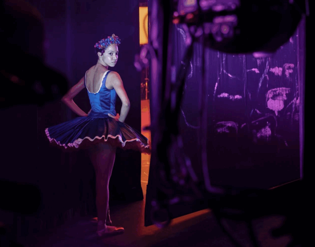Backstage in the wings, in moody purple lighting, a ballerina stands with her back to the camera but turns her head to look into the lens, hands on her hips. She wears a flower crown and a bright blue tutu.