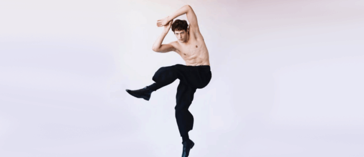 In front of a light purple and gray backdrop, Grant Gilmore jumps with his hips turned to the side, his left leg bent and lifted up. He leans with his arms bent up at the elbows, hands touching overhead. He wears black pants and dress shoes.