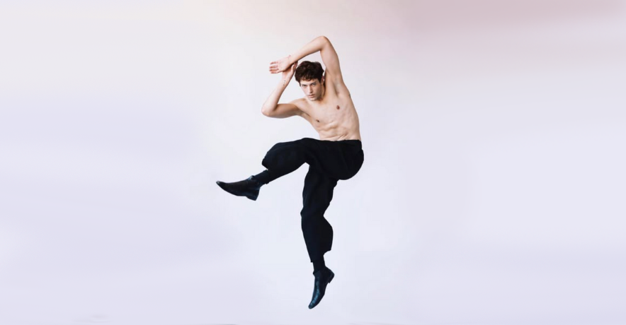 In front of a light purple and gray backdrop, Grant Gilmore jumps with his hips turned to the side, his left leg bent and lifted up. He leans with his arms bent up at the elbows, hands touching overhead. He wears black pants and dress shoes.