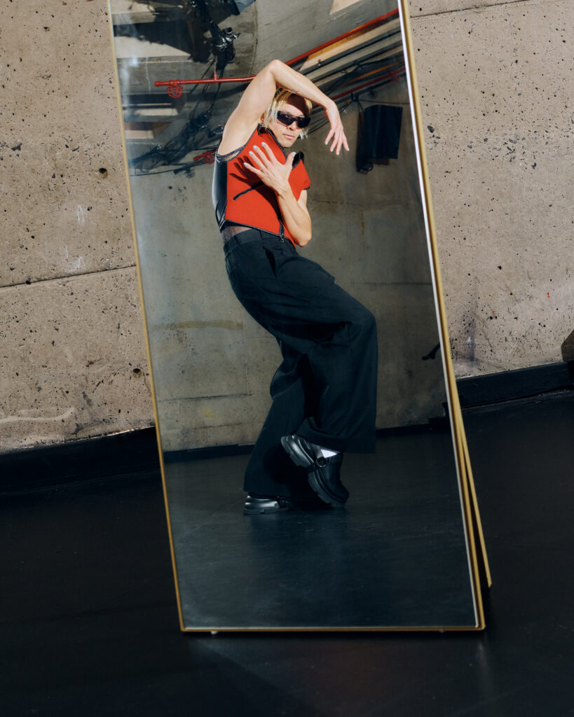 A dancer poses in a standing mirror wearing black pants, a red vest, and sunglasses. They pop one foot and wrap their arms around the torso and head.