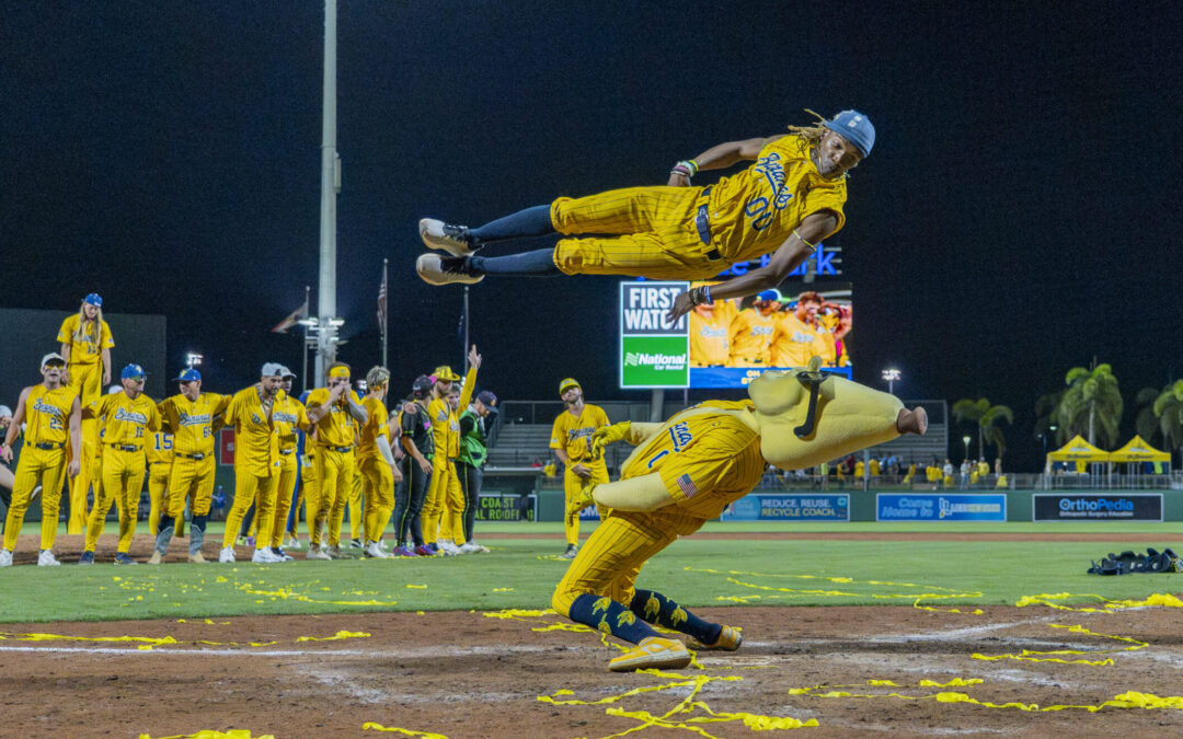 Maceo Harrison launches himself through the air while performing for a packed baseball stadium. Beneath him, a Savannah Bananas mascot hinges and leans back as Harrison jumps over him.
