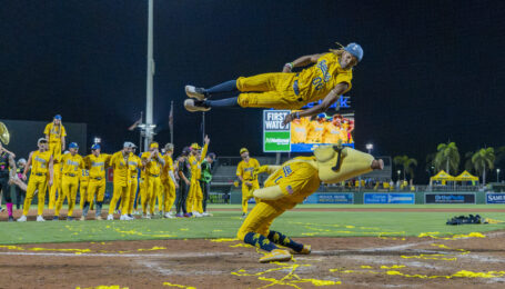 Maceo Harrison launches himself through the air while performing for a packed baseball stadium. Beneath him, a Savannah Bananas mascot hinges and leans back as Harrison jumps over him.