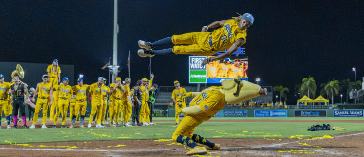 Maceo Harrison launches himself through the air while performing for a packed baseball stadium. Beneath him, a Savannah Bananas mascot hinges and leans back as Harrison jumps over him.