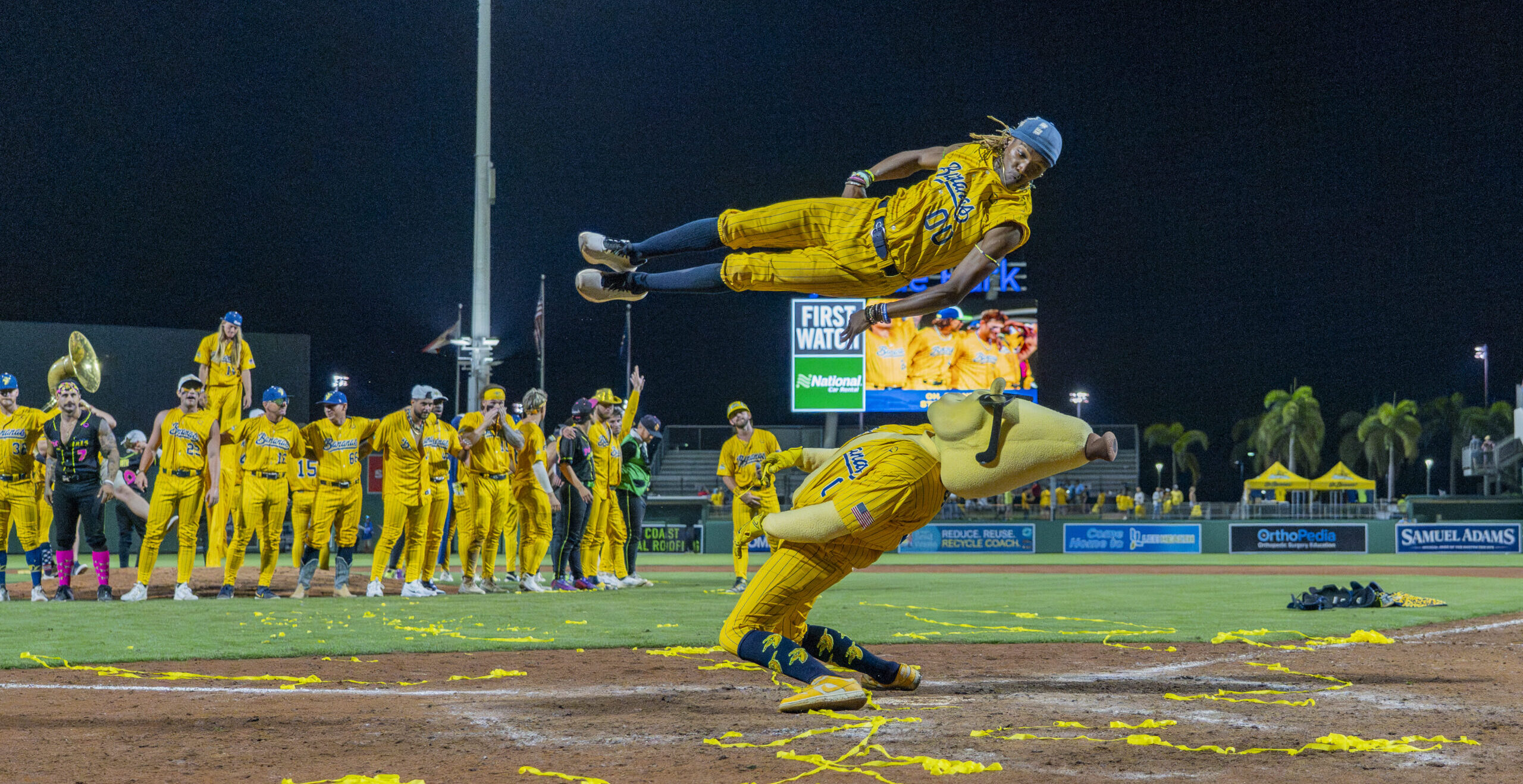 Maceo Harrison launches himself through the air while performing for a packed baseball stadium. Beneath him, a Savannah Bananas mascot hinges and leans back as Harrison jumps over him.