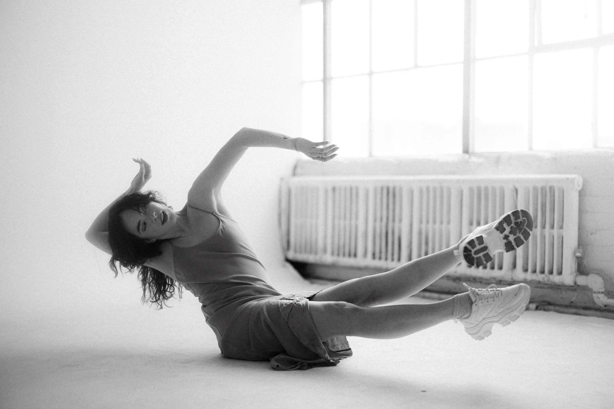 A black and white photo of a female dancer moving in a studio. She wears a simple dress and white sneakers.