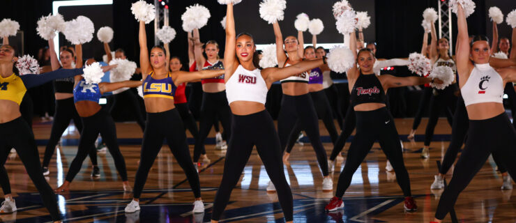 Four rows of dancers do a pom routine on a large basketball court, wearing black leggings and tops with various college logos on the front.