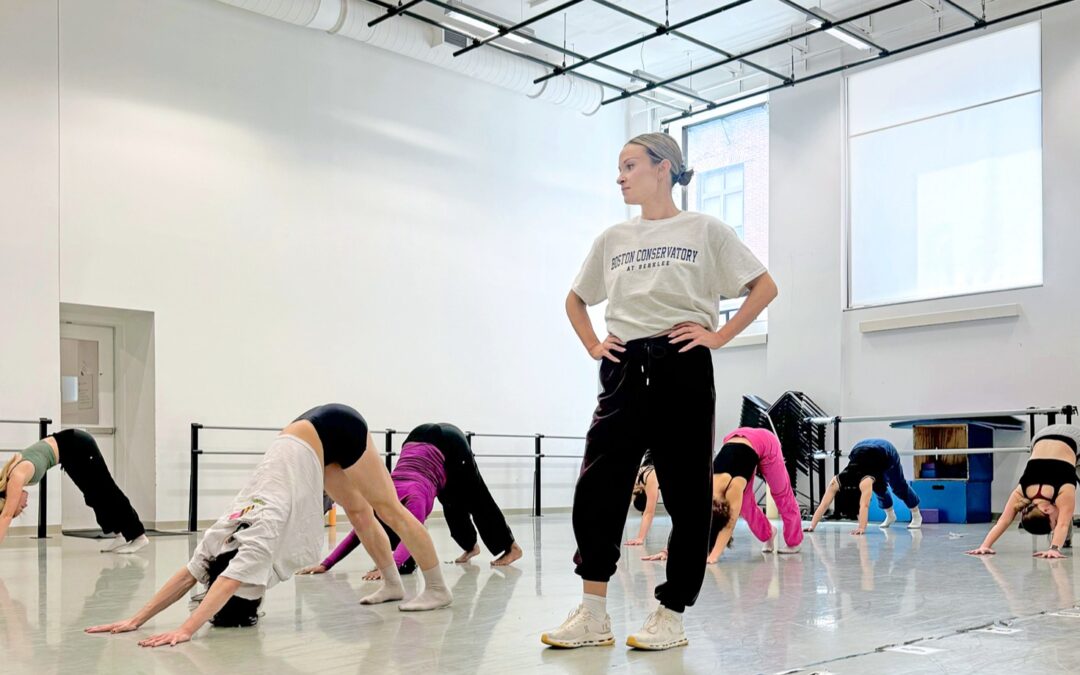 A female instructor teaching class in a studio. The students stretch in down dog while she stands and observes.
