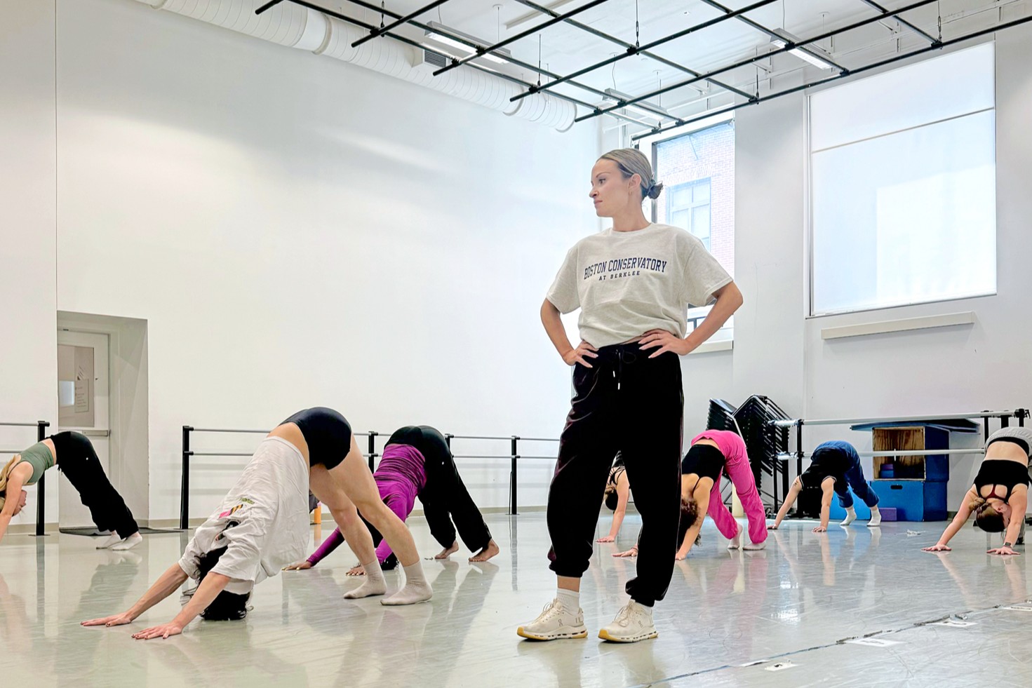 A female instructor teaching class in a studio. The students stretch in down dog while she stands and observes.