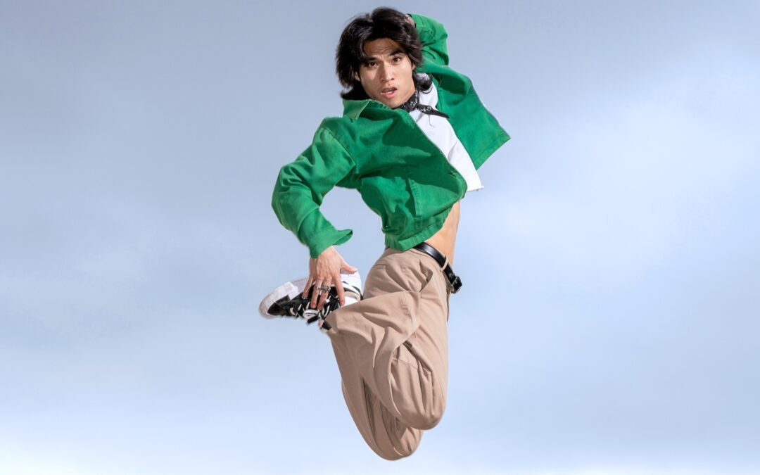Gabe De Guzman wearing a green shirt and tan pants. He jumps in the air with his legs bent.