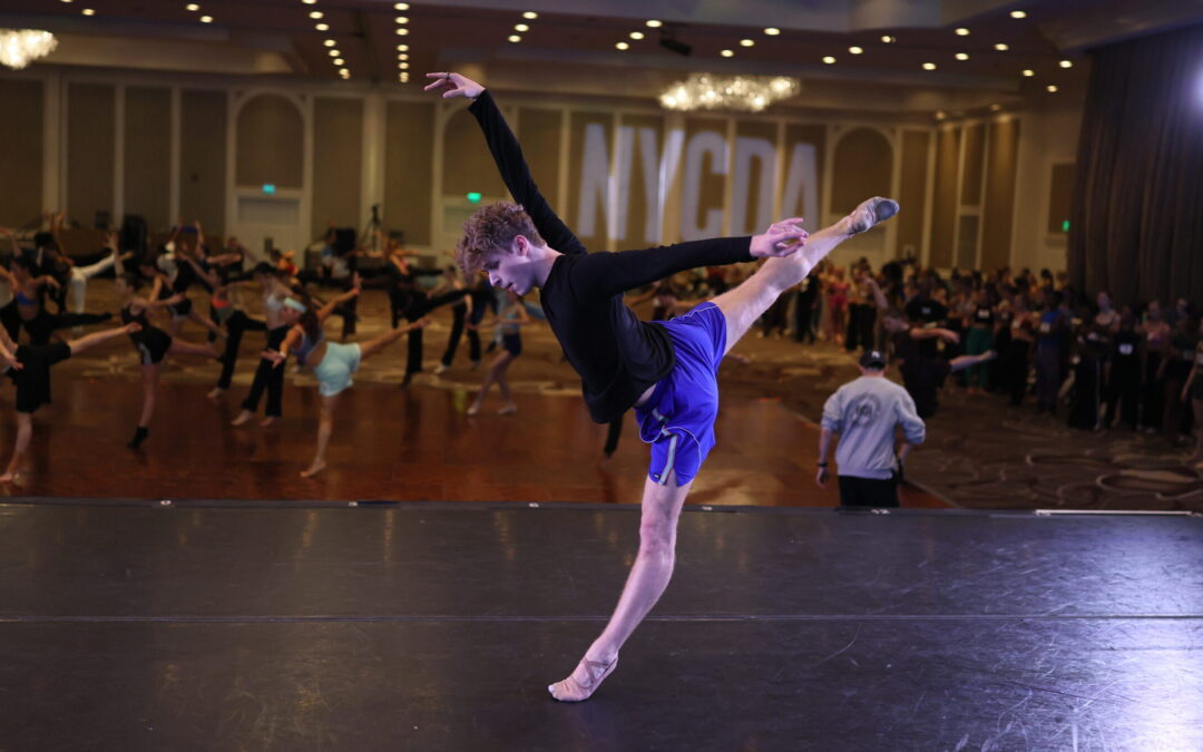 Cameron Janson sweeps one leg bag into arabesque while shifting his weight back on his supporting leg on relevé. He lifts one arm up and one back in a dramatic arabesque line, and he looks down to the floor. He dances on a stage in front of a large room of dancers at a convention.