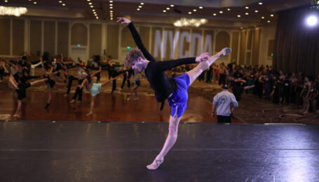 Cameron Janson sweeps one leg bag into arabesque while shifting his weight back on his supporting leg on relevé. He lifts one arm up and one back in a dramatic arabesque line, and he looks down to the floor. He dances on a stage in front of a large room of dancers at a convention.