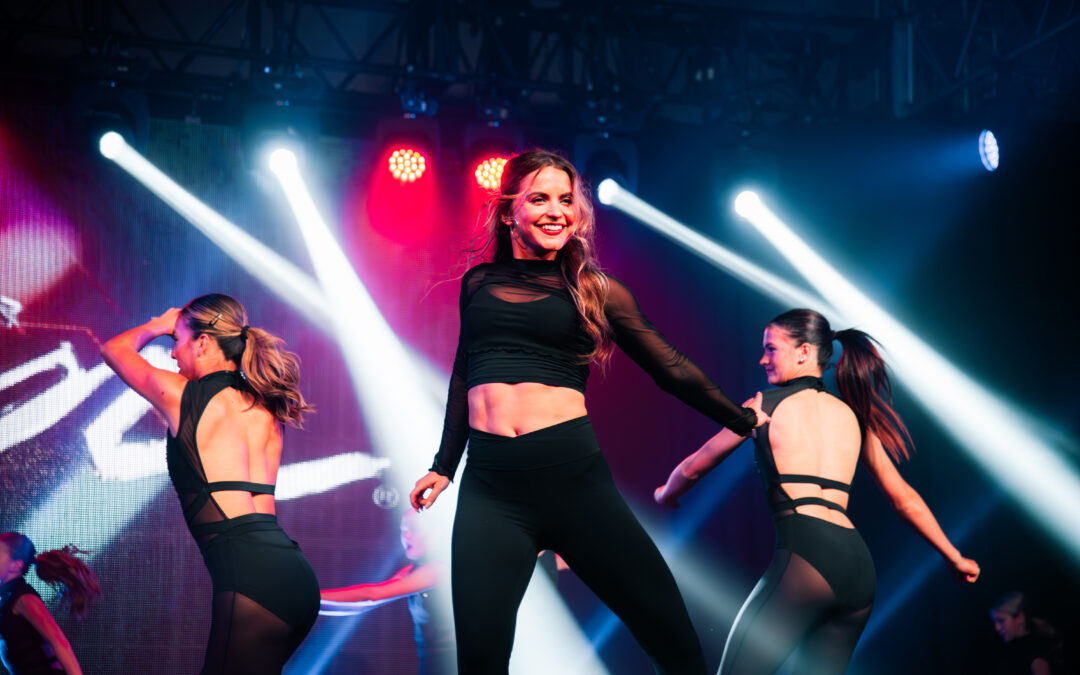 A female instructor leading on stage. She dances in front of bright lights with assistants behind her.
