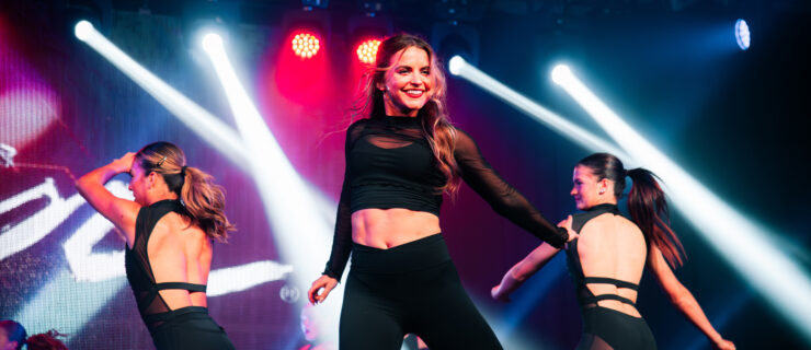 A female instructor leading on stage. She dances in front of bright lights with assistants behind her.