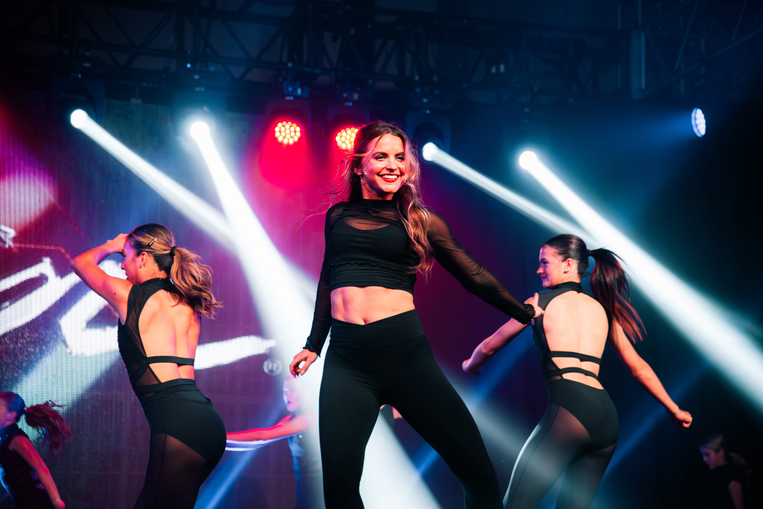 A female instructor leading on stage. She dances in front of bright lights with assistants behind her.