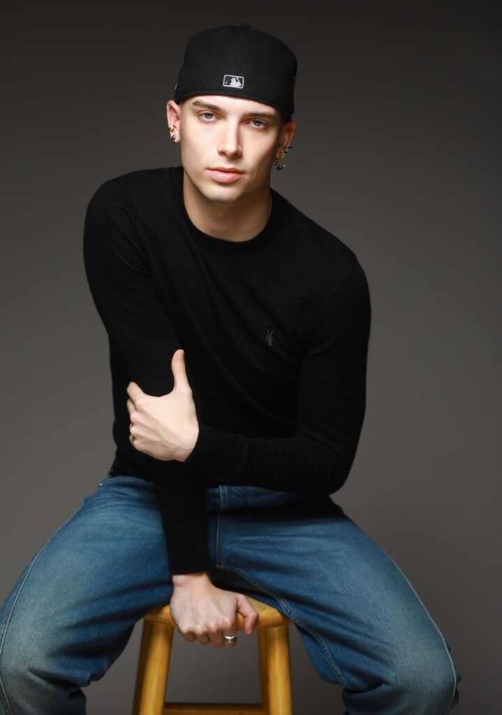 Robbie Blue, in jeans, a long-sleeved black shirt, and a backwards baseball cap, poses while sitting on a wood stool. He holds one arm and leans in with a serious expression.