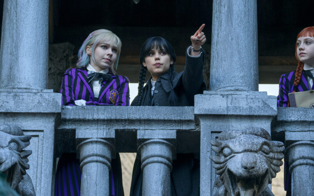 Three teenagers in striped school uniforms look over a stone balcony with gargoyles below. The middle girl points out as the other two look at her.
