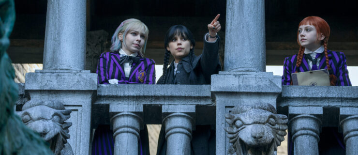 Three teenagers in striped school uniforms look over a stone balcony with gargoyles below. The middle girl points out as the other two look at her.