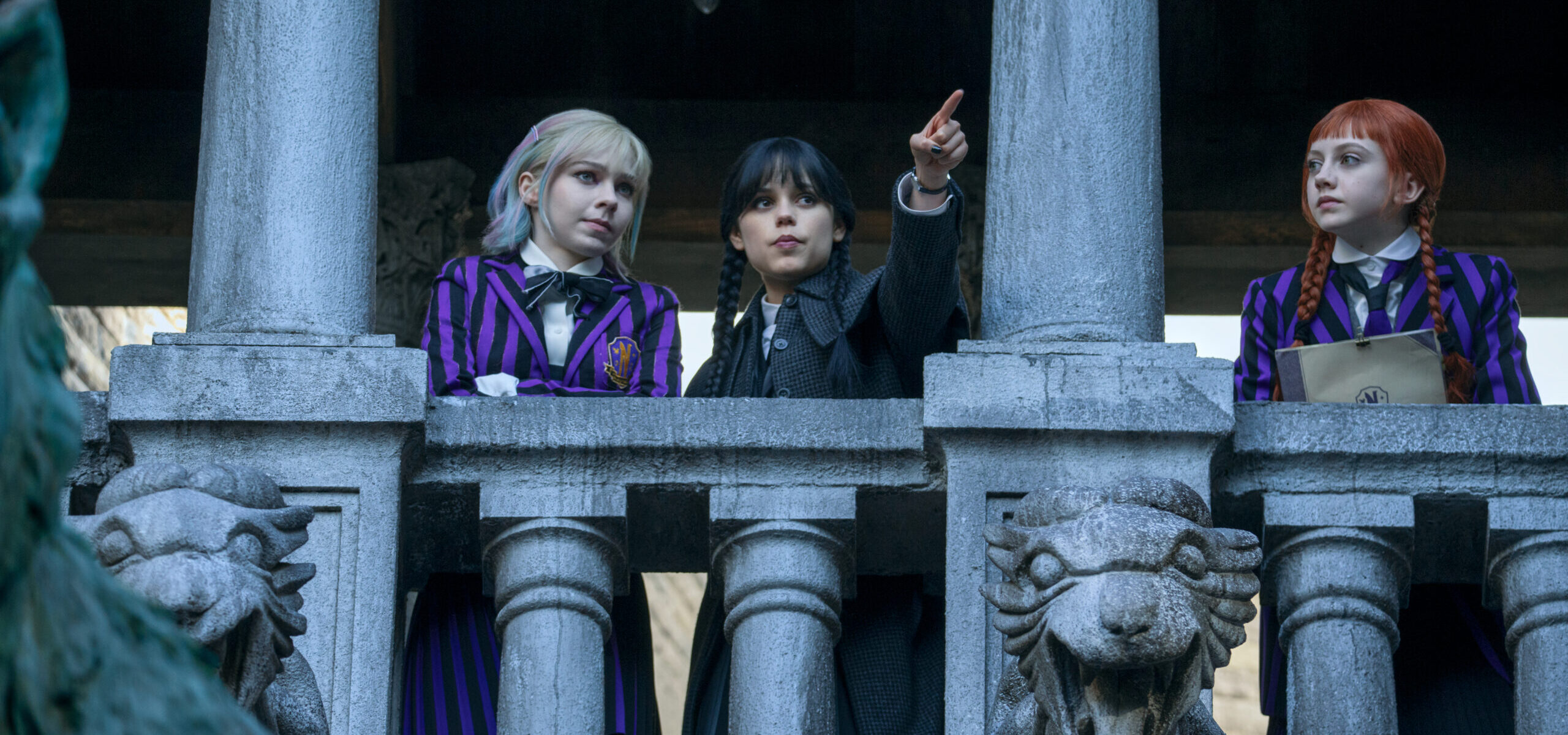 Three teenagers in striped school uniforms look over a stone balcony with gargoyles below. The middle girl points out as the other two look at her.