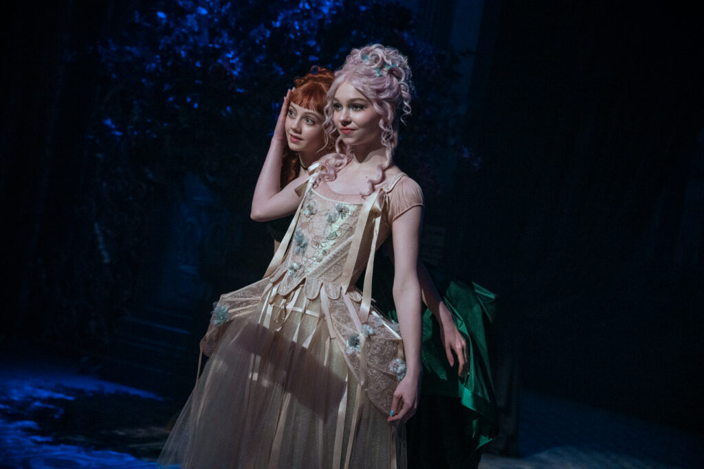 Two dancers in Venetian costumes dance in front of a dark background. The front dancer, in light pink, smiles confidently as she bends one arm up to cradle the cheek of the second dancer, who pops her head forward to rest on the first dancer's shoulder. 