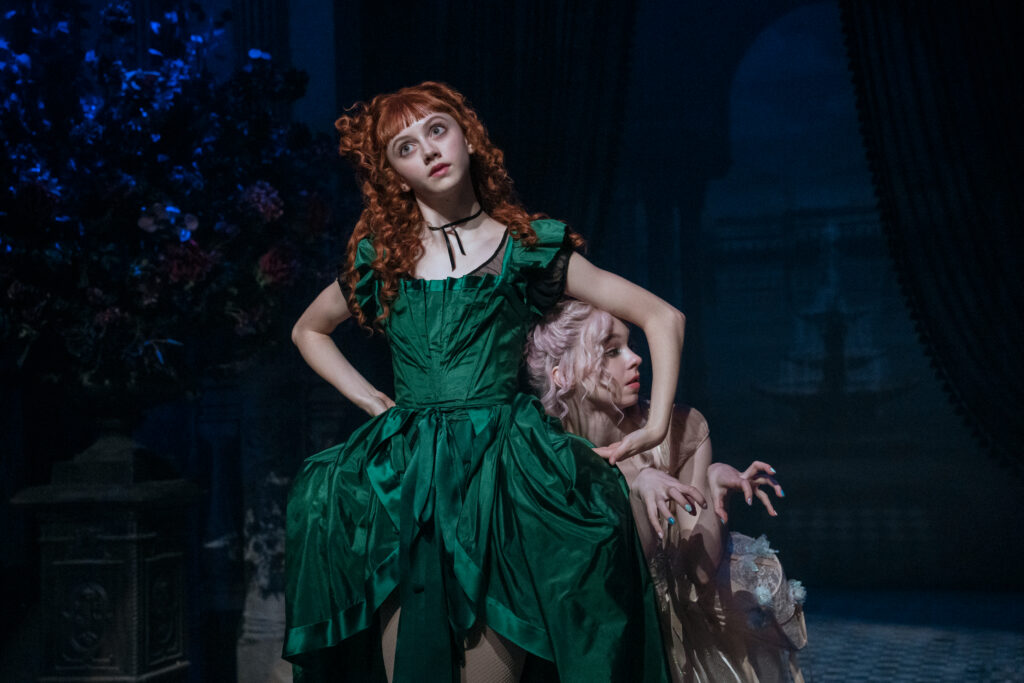 Two dancers in Venetian costumes dance in front of a dark background. The dancer in front, in green, tilts her head and bends her arms at the elbows as the second dancer, in light pink, pokes her head through the negative space.