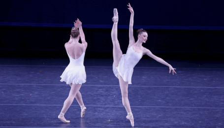 Two dancers in short white ballet dresses perform onstage. The dancer on the right battements a la seconde, arms on the diagonal as she looks out toward her bottom fingers. The dancer on the left faces upstage and crosses one foot over, with arms passing across in front of her.