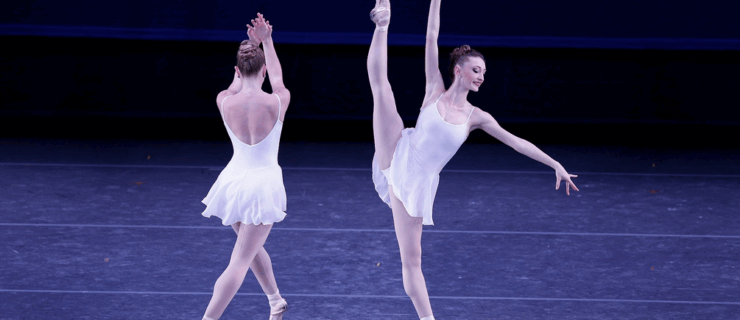 Two dancers in short white ballet dresses perform onstage. The dancer on the right battements a la seconde, arms on the diagonal as she looks out toward her bottom fingers. The dancer on the left faces upstage and crosses one foot over, with arms passing across in front of her.