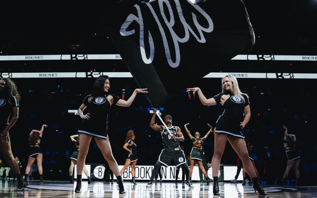 On a darkly lit basketball court, members of a dance team stand proudly across the court in sassy poses as a man waves a large Nets fan in the middle.