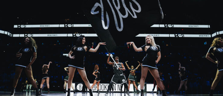 On a darkly lit basketball court, members of a dance team stand proudly across the court in sassy poses as a man waves a large Nets fan in the middle.