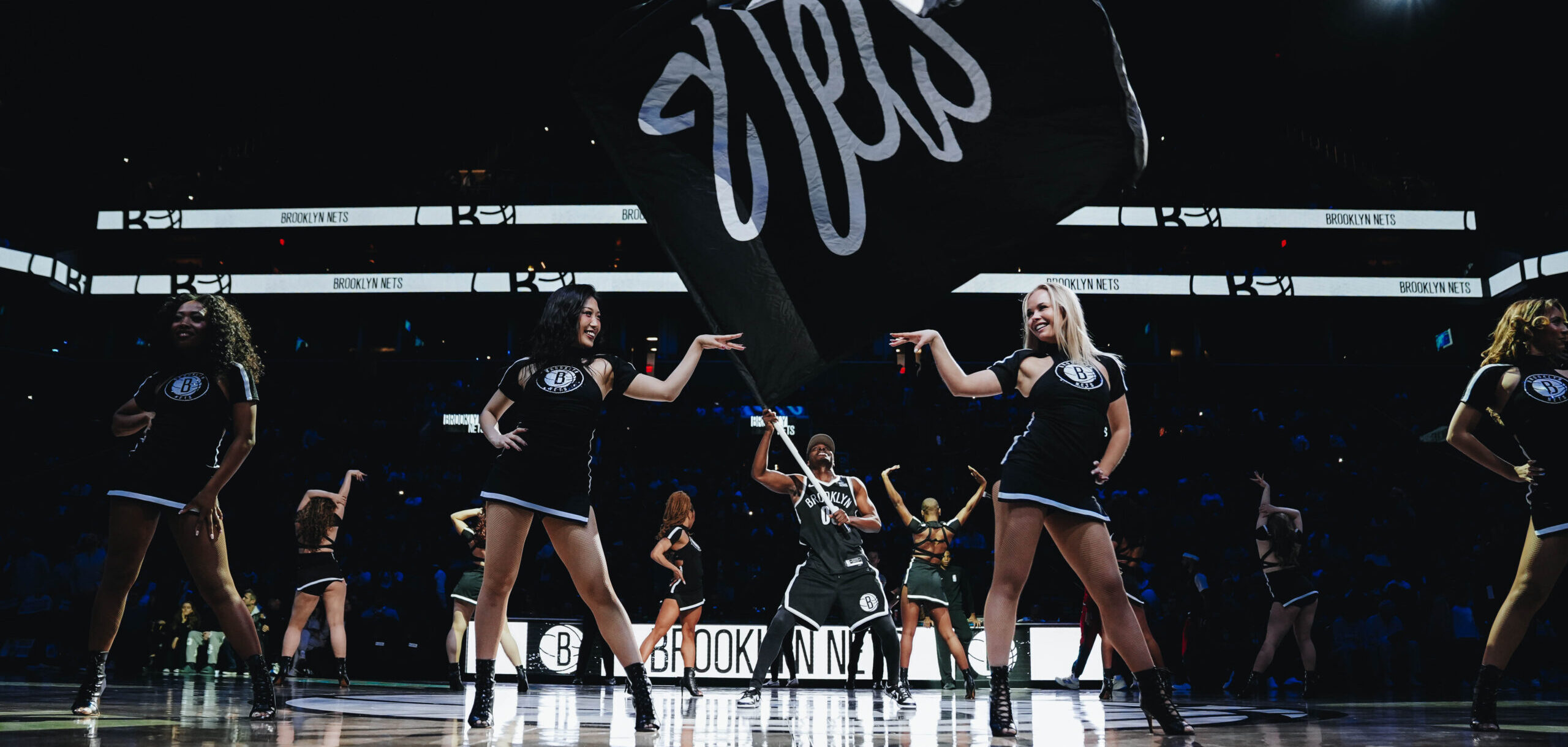On a darkly lit basketball court, members of a dance team stand proudly across the court in sassy poses as a man waves a large Nets fan in the middle.