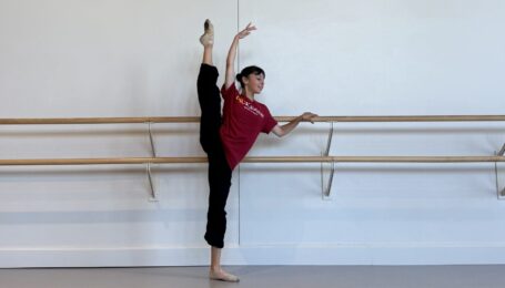 Katie Tapalaga stands with hand on the barre and her other in high fifth as she extends one leg up in a high developpe. She wears black pants and a red t-shirt.