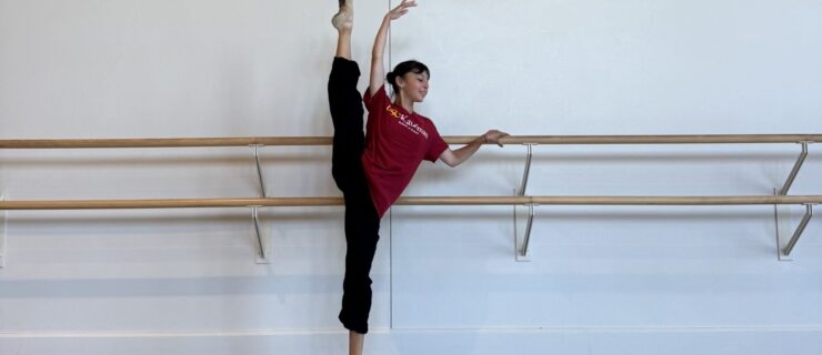 Katie Tapalaga stands with hand on the barre and her other in high fifth as she extends one leg up in a high developpe. She wears black pants and a red t-shirt.