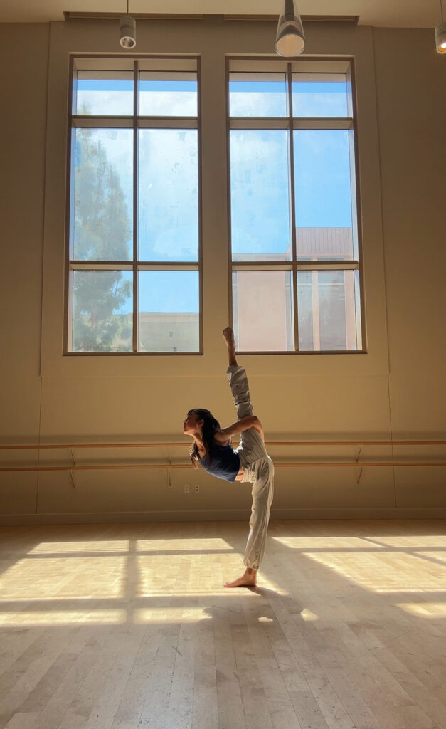 In front of a large window in a dance studio, Katie Tapalaga poses in a penché position with her arms wrapped around her back leg as she arches up to look off to the side.