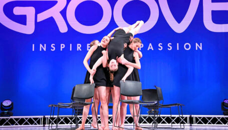 Spotlight Dance Academy dancers lifting one girl upside down on stage. She holds onto black chairs circled around.