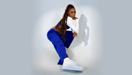 Against a white backdrop and in a spotlight, a Black dancer in blue pants, white shoes, and a long sleeve white crop top with long dark braids lunges. The photo is from the side, with her straight leg closest to the camera. Her arms are folded and one hand rests on her chin.