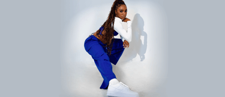 Against a white backdrop and in a spotlight, a Black dancer in blue pants, white shoes, and a long sleeve white crop top with long dark braids lunges. The photo is from the side, with her straight leg closest to the camera. Her arms are folded and one hand rests on her chin.